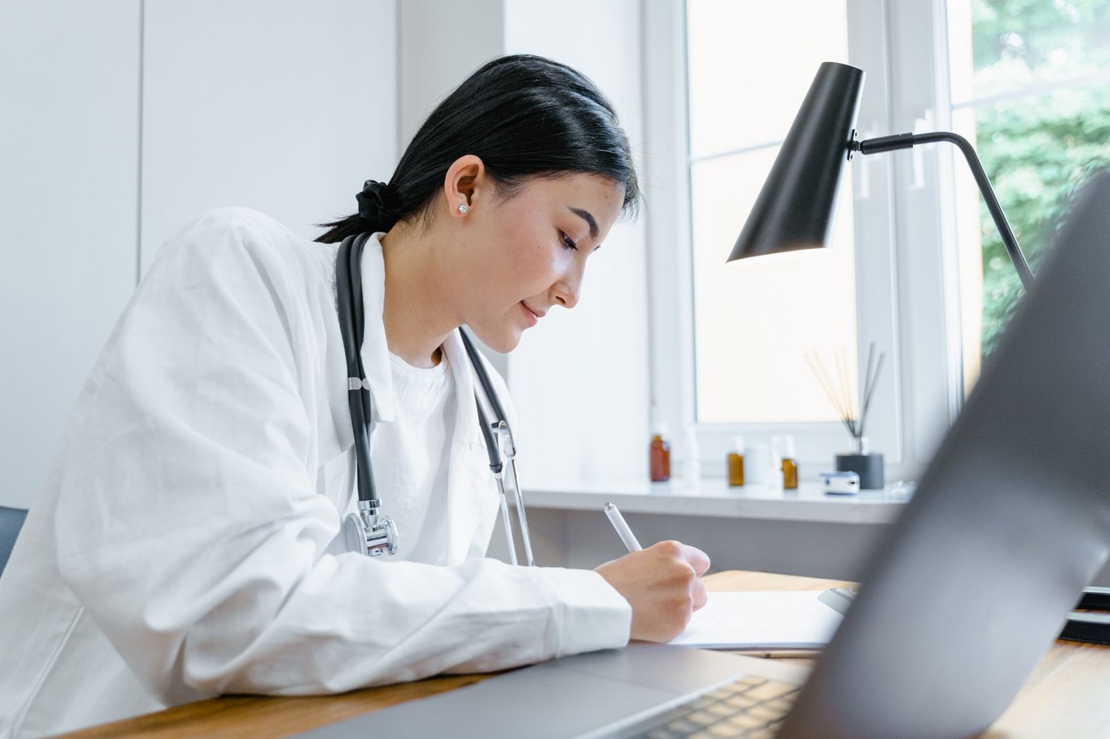 Physician in a white coat writing notes at a desk beside a laptop